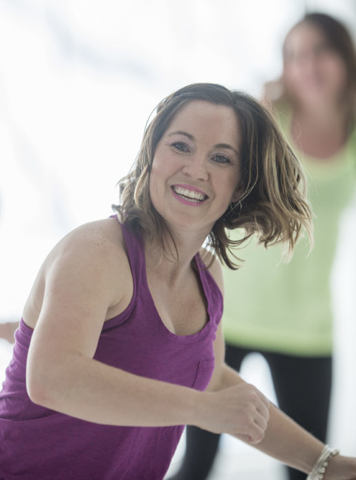 Group of women are taking an aerobic dance fitness class together at the gym. One woman is smiling and looking at the camera.